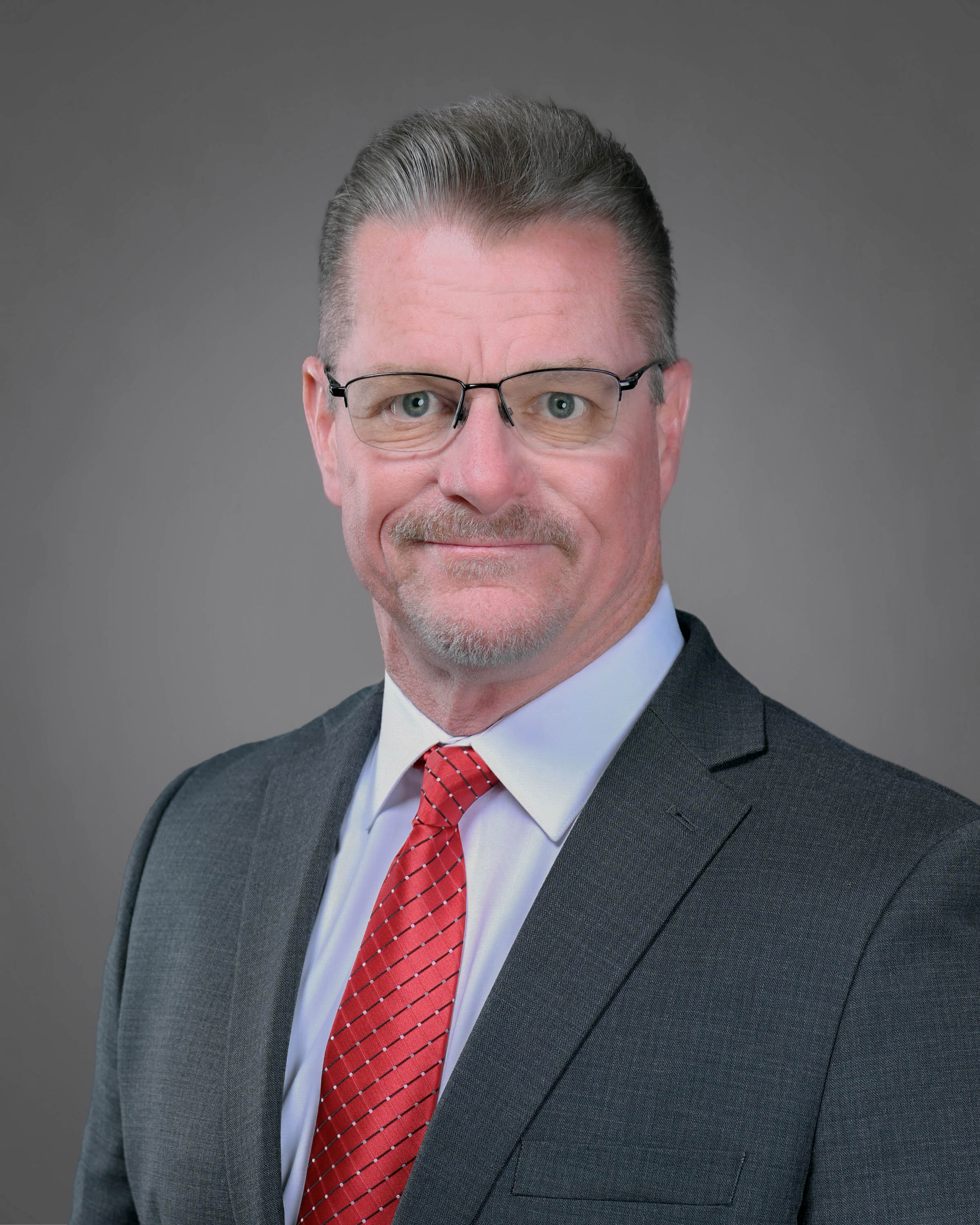 Man smiling with dark hair wearing eyeglasses in a dark suit with red tie